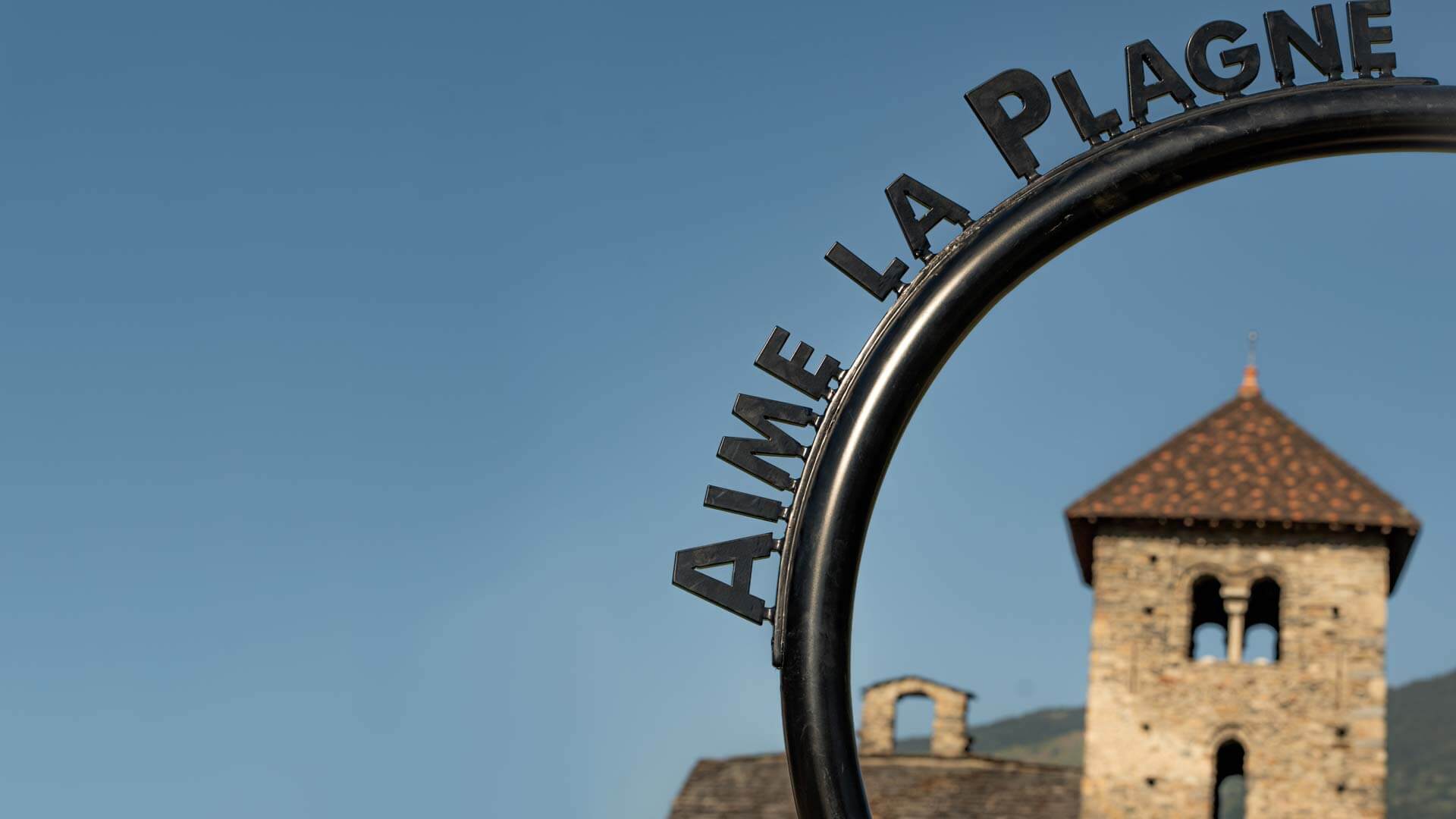 Photographie du coeur d'Aime-la-Plagne, devant la basilique, avec le nom de la commune écrit.Photographie aérienne du centre ville d'Aime-la-Plagne.