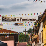 Rue principale de Longefoy avec des banderoles triangulaires colorées et une bannière avec inscrit "Pain cuit à l'ancienne, du 19 au 21 Juillet", ainsi que la chapelle de Longefoy visible en fond