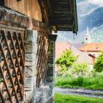 Bâtiment de type chalet, en bois et en pierre, avec un village et un clocher en arrière plan