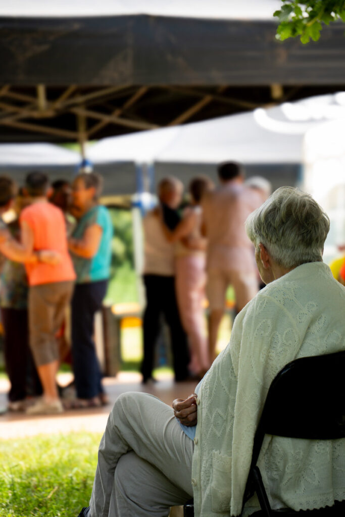 Une personne âgée est assise seule sur une chaise et observe un groupe de personnes qui socialisent en plein air sous des tentes.
