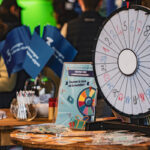 Une roue de prix colorée et des brochures sur une table lors d'un événement, avec des personnes et des drapeaux bleus en arrière-plan.