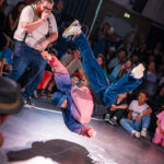 Un breakdancer tourne sur le dos sous le regard attentif d'un homme muni d'un micro et d'une foule.