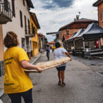 Deux personnes transportent une longue planche en bois contenant plusieurs pains dans une rue.