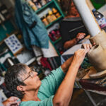 Femme âgée portant des lunettes et sculptant de l'argile dans un studio d'art avec des étagères de fournitures en arrière-plan.