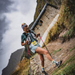 Un coureur avec des bâtons de trekking sourit lors d'une course de trail en montagne, une chapelle et un ciel nuageux en arrière-plan.