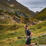 Un homme fait de la course à pied avec des bâtons de trekking dans un paysage montagneux sous un ciel nuageux.
