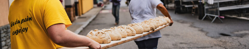 Deux personnes transportent une longue planche en bois contenant plusieurs pains dans une rue.