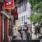 Grande rue d'Aime, étroite avec des enseignes de magasins, des bâtiments en pierre, des fleurs et une personne marchant à l'ombre.