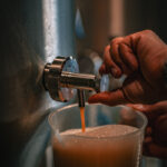 Une main verse de la bière mousseuse d'un robinet en métal dans un pichet gradué en plastique transparent.