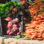 Betteraves et carottes fraîches empilées dans des caisses sur un marché, avec des légumes verts en arrière-plan.