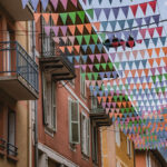 Des drapeaux triangulaires colorés sont suspendus au-dessus de la grande rue bordée de bâtiments et de balcons.