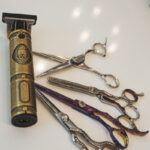 Une tondeuse électrique dorée ancienne et quatre ciseaux à cheveux ornés sur une surface blanche.