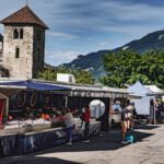 Marché en plein air avec des gens qui font leurs courses, des étals, la basilique d'Aime et des montagnes en arrière-plan sous un ciel bleu.