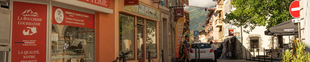 Grande rue d'Aime ensoleillée avec des magasins et des arbres bordant une rue étroite avec une vue sur les montagnes en arrière-plan.