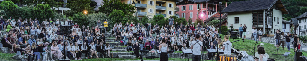Concert en plein air avec des musiciens sur scène et un large public assis sur des marches en pierre devant les maisons.