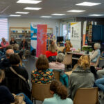 Public assis dans une librairie, écoutant une table ronde avec des orateurs à l'avant, près de banderoles colorées.
