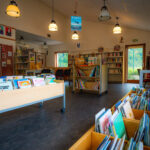 Une salle de bibliothèque colorée avec des étagères, des chariots de livres, des chaises et de grandes fenêtres laissant entrer la lumière naturelle.