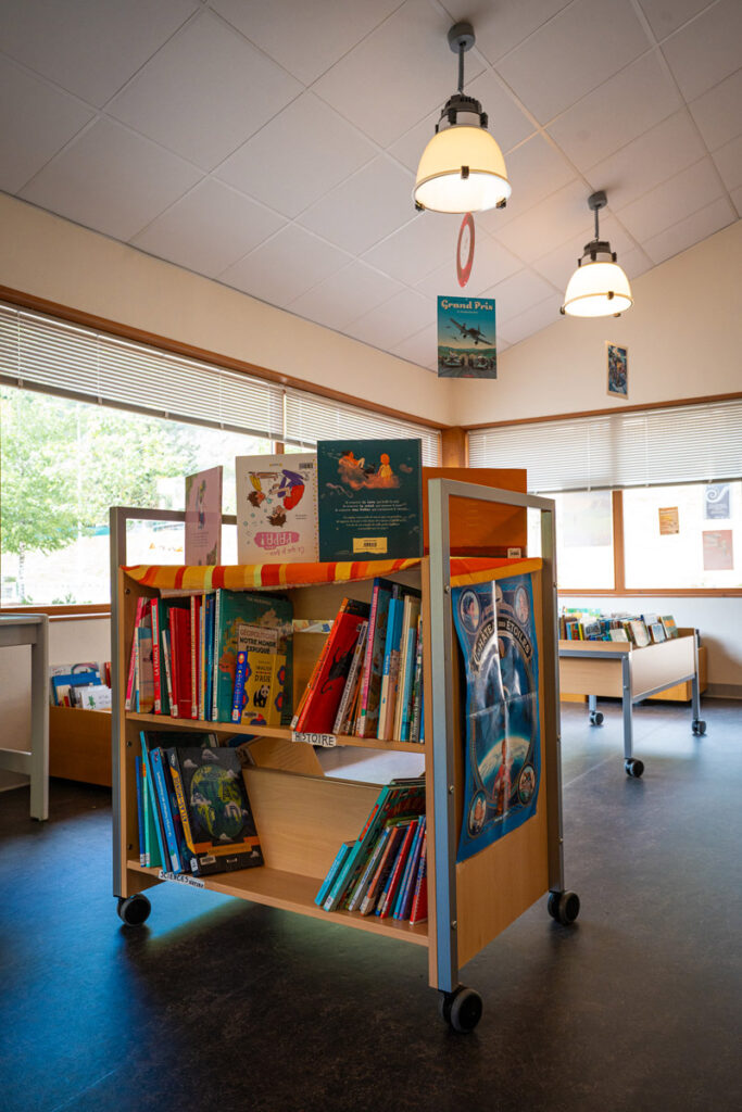 Chariot de livres rempli de livres pour enfants colorés dans une bibliothèque moderne et lumineuse avec de grandes fenêtres et des lumières suspendues.