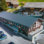 Un bâtiment alpin moderne en bois avec des accents de pierre, entouré de voitures garées et de collines verdoyantes.