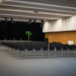 Dans un auditorium moderne, des rangées de chaises noires vides font face à une scène aux rideaux noirs et à un arbre en pot.