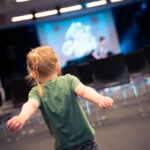 Un jeune enfant aux bras tendus se tient dans une pièce vide, face à une scène aux lumières floues.