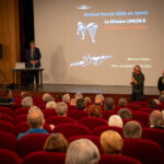 Un homme s'adresse à un public dans un cinéma ; une présentation sur la Mission Union 2 en Savoie apparaît à l'écran.