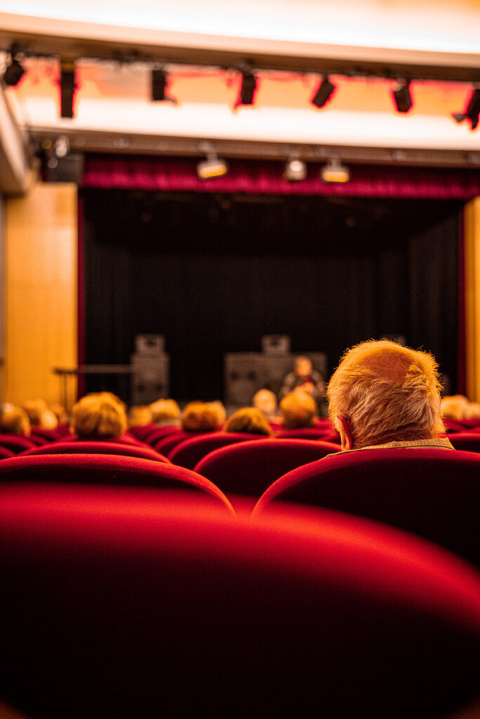 Public assis sur des chaises de théâtre rouges, face à une scène avec des haut-parleurs et un rideau noir en arrière-plan.