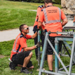 Des employés communaux des services techniques montent un échafaudage métallique en plein air, sur une pelouse, près de la basilique.