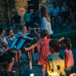 Des enfants dansent devant un groupe de musique en plein air, tandis que les gens s'assoient et regardent en soirée.