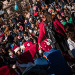 Une femme vêtue d'une veste rouge guide des enfants coiffés de bonnets de Père Noël lors d'un événement festif en plein air réunissant une foule nombreuse.