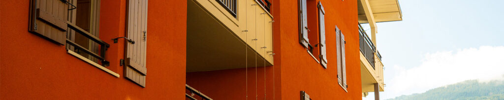 Un bâtiment moderne de couleur orange avec des volets et des balcons en bois, vu en contre-plongée dans un ciel bleu.
