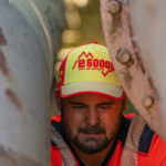 Employé de la régie des eaux, avec une casquette de la 6000D, entre deux gros tuyaux métalliques, en train de réparer