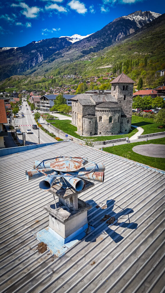 Une sirène rouillée sur le toit de la mairie surplombe la basilique, Aime et des montagnes sous un ciel bleu.