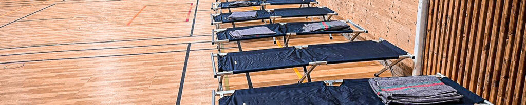 Plusieurs lits pliants avec des couvertures sont alignés contre le mur d'un gymnase au sol en bois.