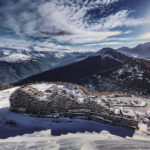 Vue aérienne d'Aime 2000 entourés de neige, de forêt et de pics lointains sous un ciel partiellement nuageux.