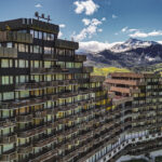 Aime 2000 : bâtiment moderne en gradins avec balcons sur fond de collines verdoyantes et de montagnes enneigées.