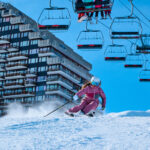 Un skieur en rose descend une piste enneigée, avec des remontées mécaniques et un grand bâtiment en arrière-plan, sous un ciel bleu.