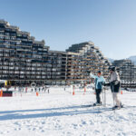Deux skieurs se tiennent sur un sol enneigé devant le paquebot des neiges à Aime 2000 avec des montagnes en arrière-plan.