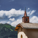 Petite église avec un clocher, entourée de montagnes vertes et d'un ciel bleu avec des nuages.