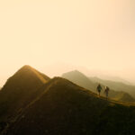 Deux personnes marchent le long d'une crête de montagne au lever du soleil, avec des sommets brumeux en arrière-plan.