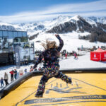 Une personne en combinaison de neige colorée saute sur un tapis gonflable dans une station de ski enneigée avec des montagnes en arrière-plan.