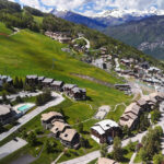 Vue aérienne de Montalbert avec chalets et routes sur des collines verdoyantes, entouré d'arbres et de lointaines montagnes enneigées.