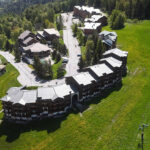Vue aérienne de bâtiments d'une station de montagne entourés de champs verts et d'arbres, avec des remontées mécaniques sur la droite.