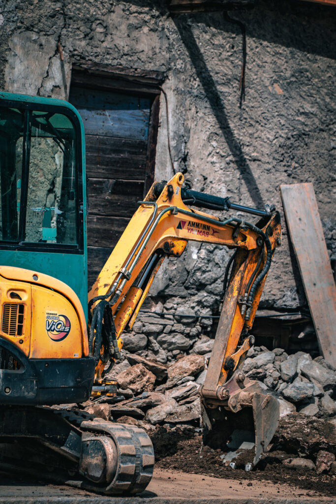 Un tractopelle devant des pierres, avec juste derrière le mur d'une ancienne bâtisse et une vieille porte en bois