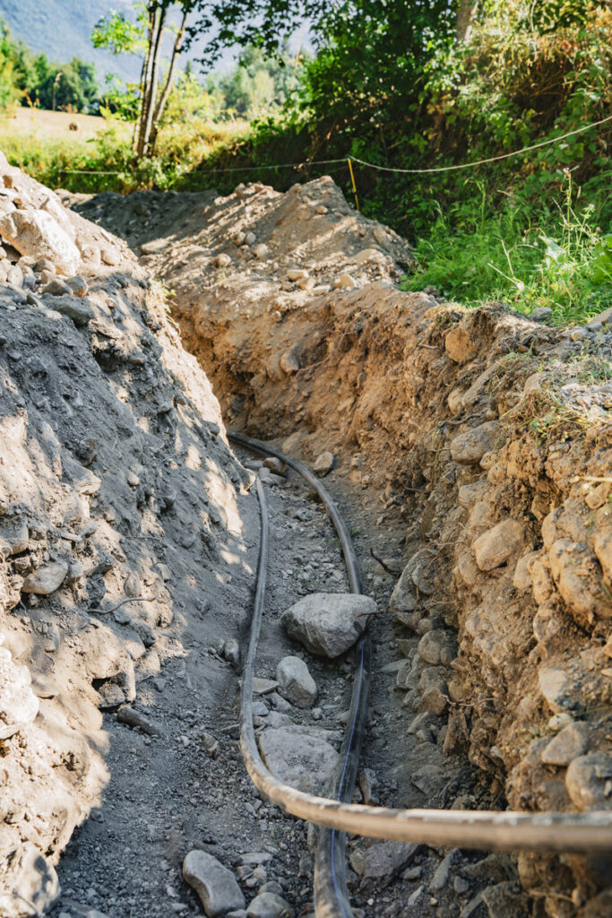 Tranchée creusée dans la terre et les cailloux pour faire passer des tuyaux, avec un champ en arrière plan