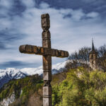 Croix en bois sur laquelle est gravée l'année 2003, entourée de montagnes, d'arbres et d'une église sous un ciel nuageux.