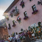 Des personnes se rassemblent à l'extérieur de la mairie de Granier dont les fenêtres sont ornées de fleurs violettes, sous un soleil éclatant.