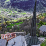 Clocher d'église avec horloge, toits, vallée verdoyante et montagnes enneigées en arrière-plan.