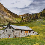 Maison en pierre sur une colline verdoyante avec des fleurs jaunes, entourée de montagnes sous un ciel nuageux.