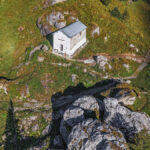 Une petite chapelle blanche au toit gris se trouve sur une colline herbeuse entourée de rochers et d'arbres.